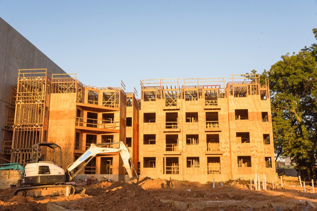 Site construction of unfinished wood framing multi-storey apartment complex, excavator on base engineering, concrete garage-wrap, pile of debris/sand. Multifamily/student housing project in Texas, USA